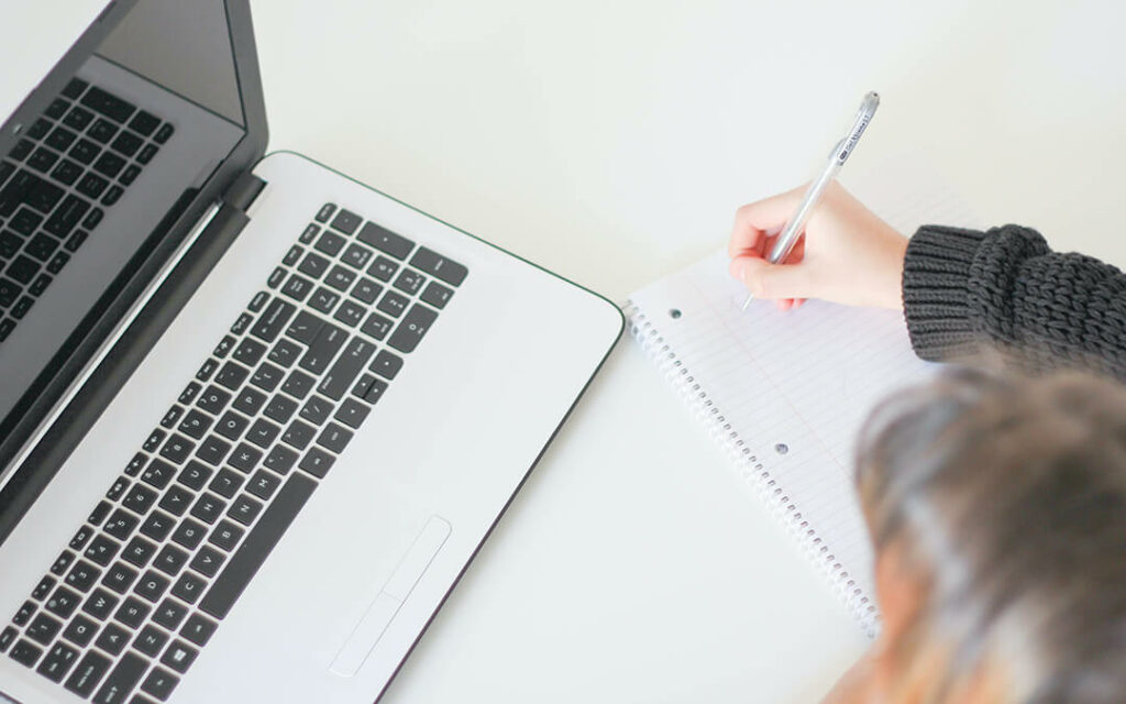 Woman writing on pad of paper next to MacBook laptop